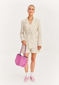 Žena v béžovém pruhovaném saku s páskem a kraťasech, držící růžovou semišovou tašku, v bílých ponožkách a růžových teniskách Adidas.