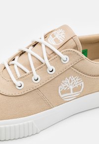 Beige canvas sneakers med vit snörning, med en vit trädlogga på sidan och en texturerad gummisula.
