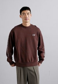 Καφέ φούτερ με ριγωτό λαιμό και μανίκια, διαθέτει μικρό λογότυπο στο στήθος και κείμενο κατά μήκος του δεξιού μανικιού. Φορεμένο με γκρι παντελόνι.