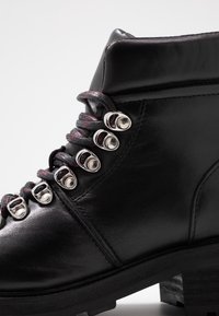 Zwarte leren enkelboot met een gevoerde kraag, robuuste ronde veters met rode accenten en zes paar zilverkleurige metalen vetersluitingen.