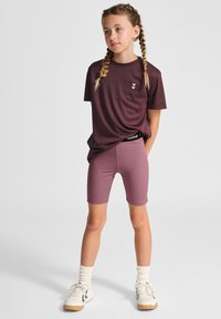 Bordeauxkleurig T-shirt met korte mouwen en logo, gecombineerd met bijpassende mauve fietsbroek. Heeft een ontspannen pasvorm. Witte sneakers en sokken tot de enkel maken de look compleet.