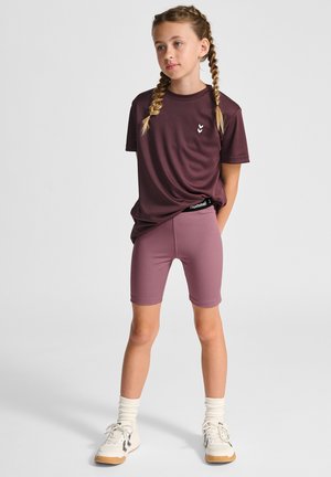 Bordeauxkleurig T-shirt met korte mouwen en logo, gecombineerd met bijpassende mauve fietsbroek. Heeft een ontspannen pasvorm. Witte sneakers en sokken tot de enkel maken de look compleet.
