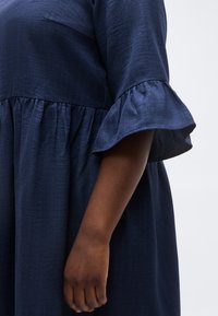 Σκούρο δέρμα χεριού που κρέμεται δίπλα σε μπλε-ναυτικό φόρεμα με σούρα στη μέση και μανίκια που φουσκώνουν μήκους μέχρι τον αγκώνα, πάνω σε ανοιχτόφωρο φόντο.