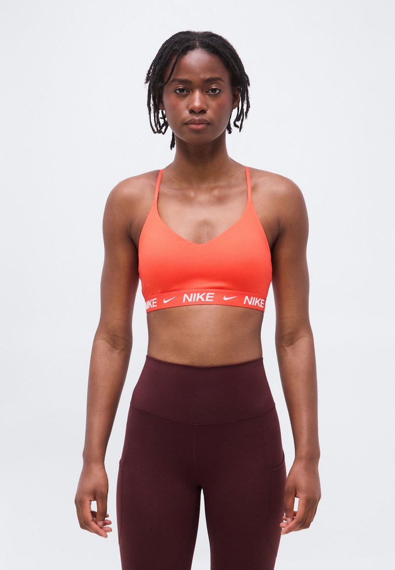 Donna atletica con dreadlocks corti, che indossa un reggiseno sportivo Nike arancione e leggings aderenti bordeaux a vita alta, in piedi davanti a uno sfondo semplice.