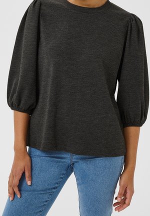 Haut gris foncé texturé avec manches bouffantes jusqu'au coude, associé à un jean en denim bleu sur une femme avec un fond uni.