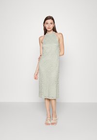 Vestido midi de renda verde claro com gola alta e padrão floral; apresenta alças e cai abaixo dos joelhos. Combinado com sandálias simples.
