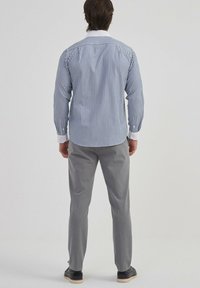 Chemise rayée bleue et blanche à manches longues avec des poignets blancs contrastants, associée à un pantalon gris et des chaussures noires. Vue de dos.