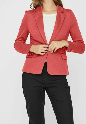Femme boutonnant un blazer corail sur un top blanc, portant un pantalon noir, vue du cou aux hanches sur un fond uni.
