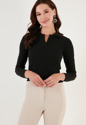 Black ribbed long-sleeve top with a collar and a front slit, paired with beige fitted pants. Earrings feature a circular pattern in black and gold.
