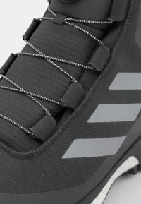Czarne sportowe buty z textured górą, odblaskowymi srebrnymi akcentami i elastycznymi sznurówkami zabezpieczonymi szarymi złączkami.