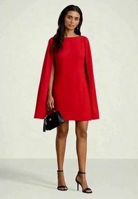 Kvinne iført en rød kappekjole som holder en liten svart håndveske, står med svarte høyhælte sandaler foran en ensfarget bakgrunn.