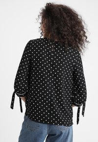 Černá halenka s puntíky, tříčtvrteční rukávy, detaily vázání na manžetách a klíčovým otvorem na zádech. Měkká látka s hladkou texturou.