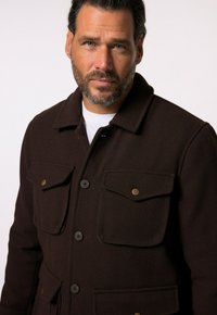 Chaqueta de lana marrón oscuro con cuello, cuatro bolsillos frontales, botones negros y un sutil detalle de textura en la tela.