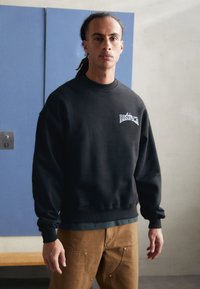 Black sweatshirt with a round neck and ribbed cuffs. Features a graphic logo on the chest. Worn with light brown pants.