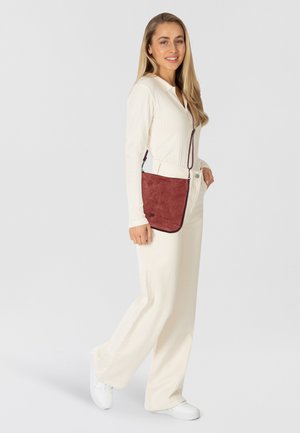 Sac bandoulière en daim rouge avec une texture lisse, doté d'une longue sangle et d'un logo discret. Le modèle porte des vêtements crème et des baskets blanches.