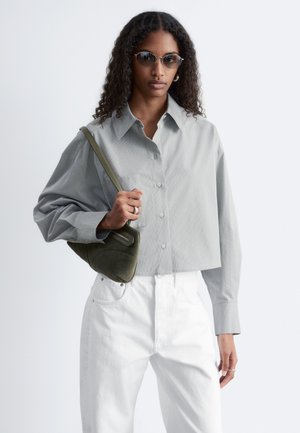 Kobieta o kręconych włosach, nosząca okulary przeciwsłoneczne, jasnoszarą krótko skrojoną koszulę zapinaną na guziki, białe spodnie i trzymająca oliwkowo-zieloną torbę na ramię.