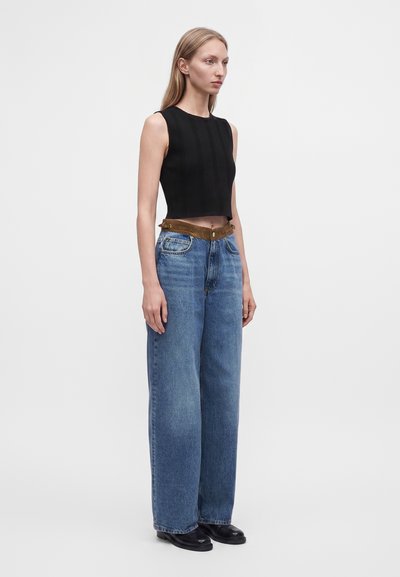 Femme aux cheveux blonds raides portant un crop top noir sans manches, un jean bleu ample avec une ceinture marron et des chaussures noires, debout de profil.