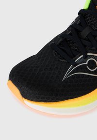 Zapatilla deportiva negra con parte superior de malla, que presenta un logo negro y plateado, acentos naranjas en la suela y una superficie texturizada.