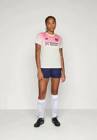 Woman standing wearing a pink and white soccer jersey, navy shorts, white knee-high socks, and black cleats against a plain background.