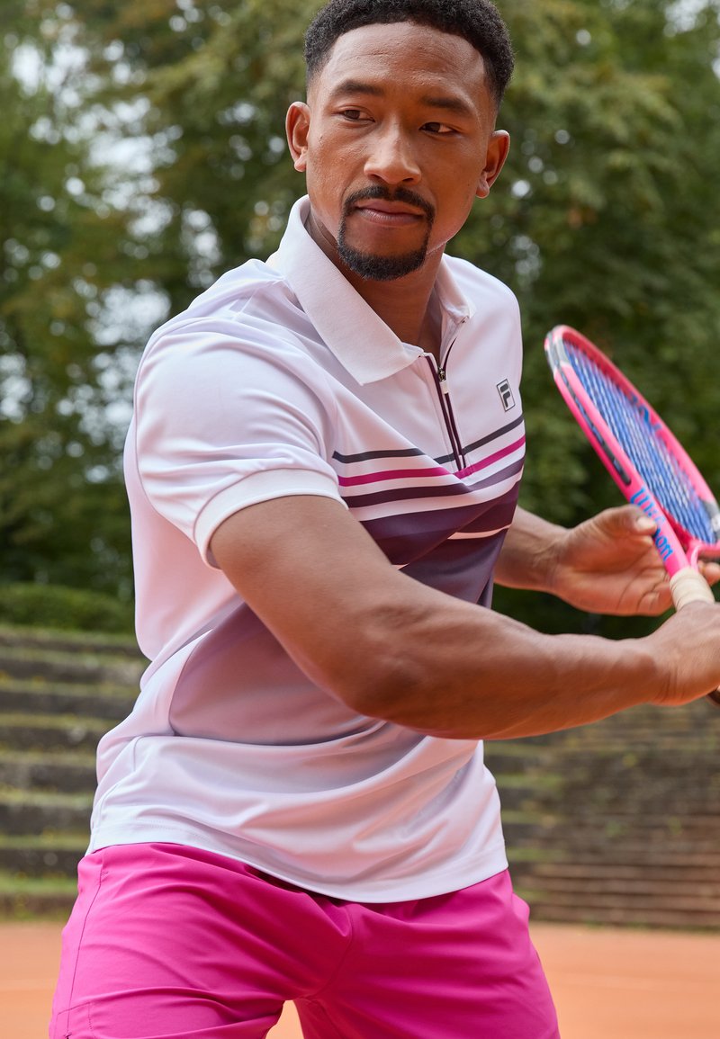 Polo shirt blanc et violet à rayures associé à un short rose, tenant une raquette de tennis. Accent sur les vêtements de sport et l'action.