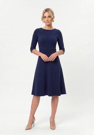 Femme portant une robe bleu marine au genou avec des manches trois-quarts et des talons hauts couleur nude, debout devant un fond blanc uni.