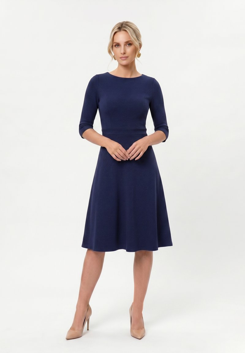 Femme portant une robe bleu marine au genou avec des manches trois-quarts et des talons hauts couleur nude, debout devant un fond blanc uni.