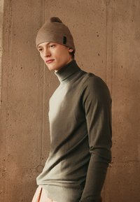Suéter de gola alta em verde oliva claro com nervuras, combinado com um gorro bege também com nervuras. Textura suave, design ajustado e estilo minimalista.