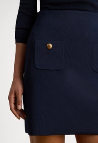 Σκούρο μπλε ναυτικό, φούστα με ριμπ υφή που διαθέτει δύο μπροστινές τσέπες με χρυσά κουμπιά. Κοντινή λήψη, χέρι ακουμπισμένο στην επιφάνεια της τσέπης.