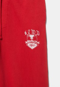 Czerwona tkanina z białą grafiką przedstawiającą orła, napisami „STWD”, „Forever Authentic” oraz „1991” na emblematycznym tarczy.