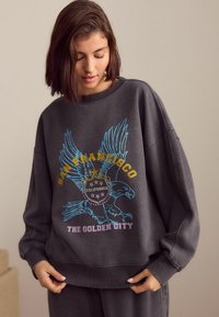 Šedá oversized mikina s barevným vyšíváním orla a textem "San Francisco, Kalifornie, Zlaté město." Měkká textura.