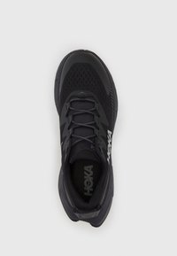 Musta urheilukenkä, jossa on verkkopinta, pehmustettu kaulus ja nauhakiinnitys. Sivussa HOKA-brändäys ja teksturoitu kumipohja.