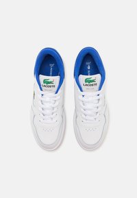 Valged tossud siniste ääristega, mille külje peal on roheline Lacoste'i logo, valged paelad ja perforatsioonid varbapiirkonnas.