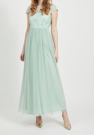 Kobieta w jasnooliwkowej, długiej sukni z kwiatową koronkową górą i przezroczystymi, krótkimi rękawkami, w zestawieniu ze złotymi szpilkami z ostrym noskiem.