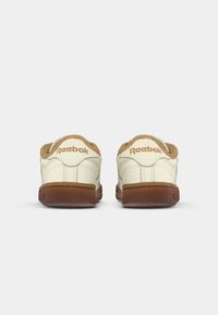 Tenisky Reebok s béžovými akcentmi, krémovým koženým zvrškom, polstrovaným golierom a hnedou gumovou podrážkou. Zadný pohľad ukazuje logo a detaily prešívania.