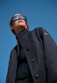 Veste légère noire avec fermeture à boutons, col texturé et boutons brillants; portée sur un haut en maille noire devant un fond de ciel bleu.