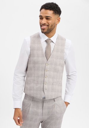 Mann lächelnd, trägt eine hellgraue karierte Weste und passende Hose mit weißem Hemd und grauer Krawatte, Hände in den Taschen vor einfarbigem Hintergrund.