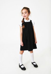 Czarna bawełniana sukienka ogrodniczka z dwiema kieszeniami, noszona na białej koszuli z kołnierzykiem. Stylizowana z białymi skarpetkami do kolan i czarnymi butami.