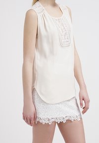 Blouse sans manches crème avec un panneau en dentelle détaillé à l'avant et un ourlet festonné. Associée à un short en dentelle blanche avec un motif floral.