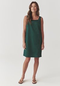 Green sleeveless dress with a square neckline, made of smooth fabric, falling to mid-thigh, paired with simple brown sandals.