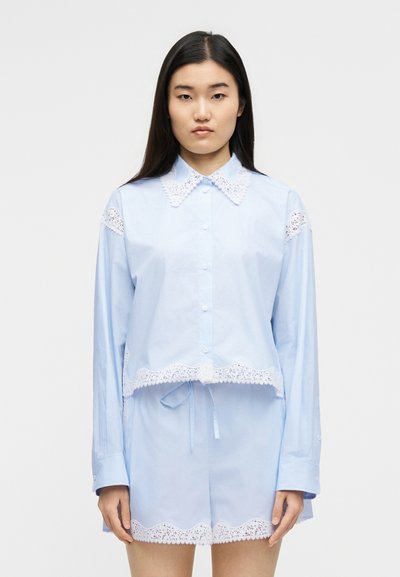 Γυναίκα που φορά γαλάζιο πουκάμισο με κουμπιά και ταιριαστό σορτς με λευκή δαντέλα στη χαλίστρα, στέκεται μπροστά σε απλό φόντο.