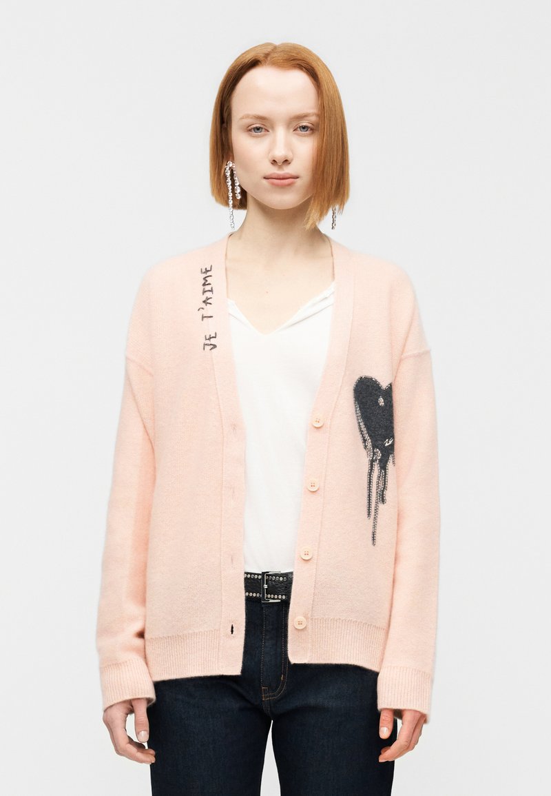 Ροζ πλεκτή ζακέτα με σχέδιο καρδιάς στη δεξιά πλευρά, με το κείμενο "Je t'aime". Τα κουμπιά είναι ανοιχτόχρωμα και η υφή είναι απαλή.