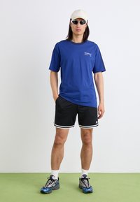 T-shirt en coton bleu à manches courtes avec un logo, associé à un short en mesh noir avec des bordures blanches et des baskets bleues. Les accessoires comprennent une casquette et des lunettes de soleil.