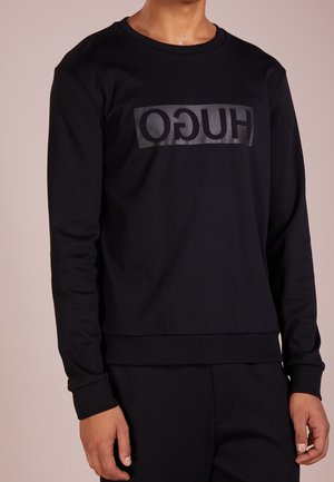 Person trägt schwarzen Sweatshirt mit langen Ärmeln und einem dunklen, glänzenden "HUGO"-Logo, das rückwärts über die Brust gedruckt ist, kombiniert mit einer schwarzen Hose.