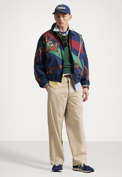 Veste bomber colorée à motifs avec design équestre, pull en maille vert, pantalon beige et baskets navy. Casquette avec lettrage.