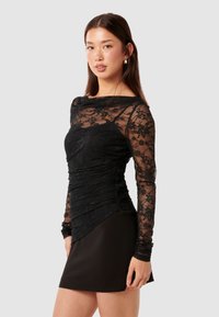 Μαύρη δαντελένια μπλούζα με μακριά μανίκια, με ρυτιδωτή υφή, εφαρμοστό κορσέ και απλωτό στρίφωμα, συνδυασμένη με μια μονόχρωμη μαύρη μίνι φούστα.