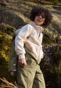 Fleece-Zip-Jacke in Creme mit strukturiertem Stoff, unter einer grünen Oversized-Jacke geschichtet. Olivgrüne Hose mit sichtbar detailierten Reißverschluss.