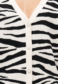 Ζακέτα με λαιμόκοψη σε σχήμα V με ασπρόμαυρες ρίγες ζέβρας, διακοσμημένη με τρία κουμπιά με μαργαριτάρι στη μέση στην μπροστινή πλευρά.