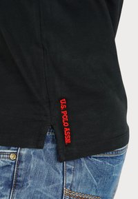 Fekete póló oldalhasítékkal, az alján vörös hímzéssel, amelyen ez áll: "U.S. POLO ASSN." Kék farmernadrág felett viselve.