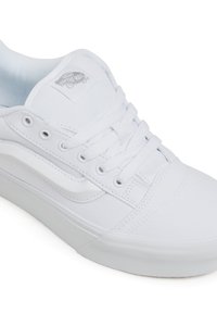 White canvas sneaker with a rounded toe, flat laces, and a rubber sole. Features the Vans logo on the tongue and side stripe design.
