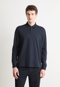 Tamsiai mėlynas polo marškinėliai ilgomis rankovėmis, pagaminti iš medvilnės. Yra mygtukų prasegimas ir siuvinėtas logotipas ant krūtinės. Lygus paviršius ir pritaikyta fasonas.